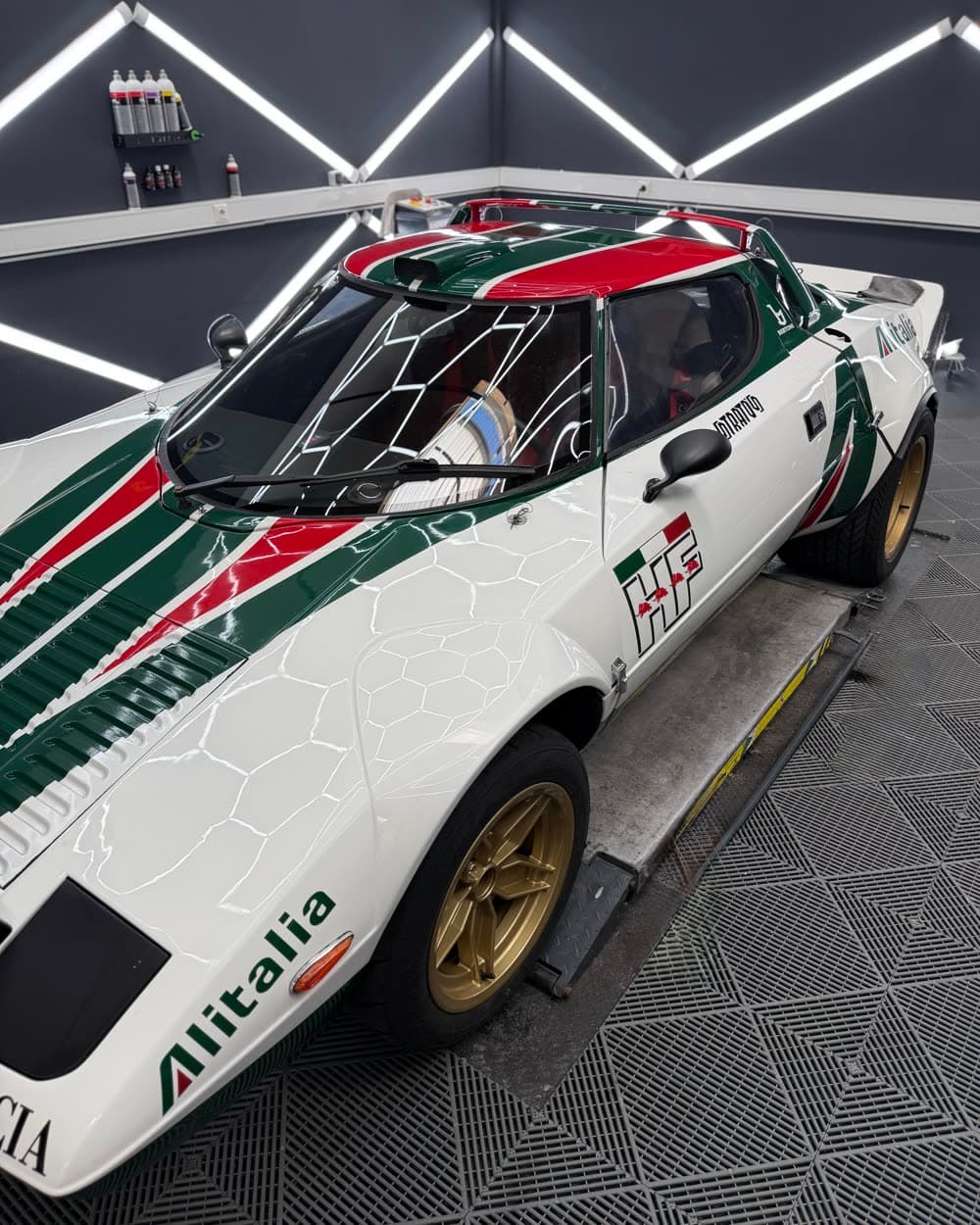 Weißer Lancia im Alitalia-Rallyedesign auf der Hebebühne, schräg von der Seite; goldene Felgen und der Schriftzug Alitalia am Schweller.