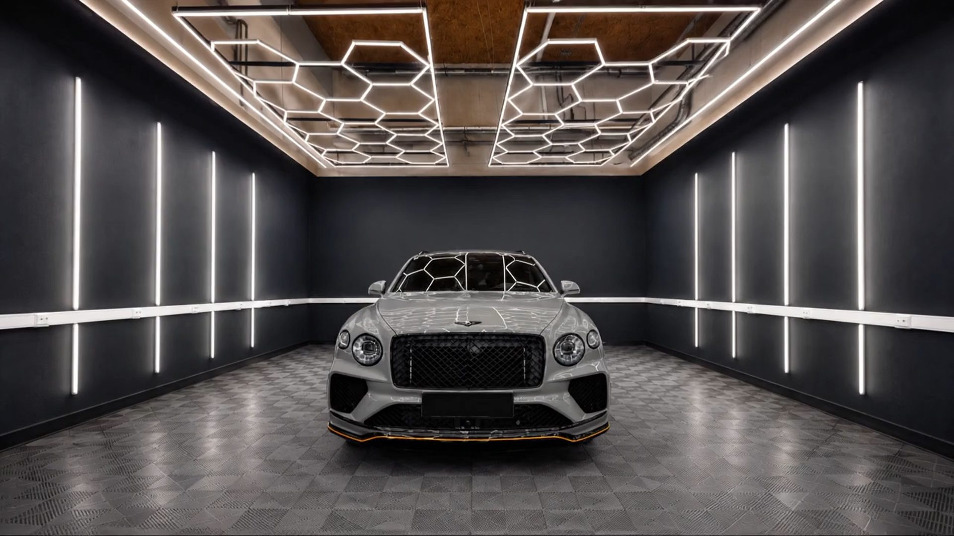 Grauer Bentley frontal in der Aufbereitungshalle: über dem Wagen ein Wabenraster aus Leuchtstreifen an der Decke, an den dunklen Wänden senkrechte Lichtleisten, darunter der graue Fliesenboden.